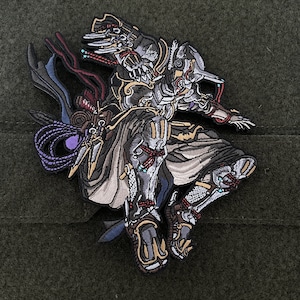 May include: Embroidered patch featuring a detailed warrior figure in a dynamic pose. The patch showcases a palette of gray, black, gold, and red, with intricate stitching and design elements. The warrior is adorned with armor and flowing fabrics, creating a sense of movement.