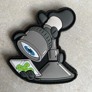 May include: A black and grey microscope patch with a green and white creature on a slide. The microscope has a large eye lens and a black and white handle.