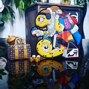 Może przedstawiać: Kreskówkowa postać jaszczurki z torbami na zakupy, kartami kredytowymi i konsolą do gier. Jaszczurka jest żółta z brązowymi i niebieskimi akcentami. Widoczna jest również mała torebka ze złotymi akcentami i złotym topem.