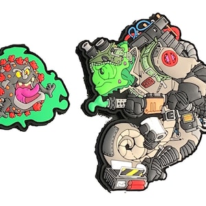 May include: Two colourful rubber charms. One is a cartoon virus with a green outline. The other is a green alien in a Ghostbusters-style outfit with a proton pack and other equipment. The charms are detailed and vibrant.
