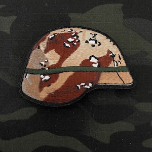 Puede incluir: Un parche de casco de camuflaje marrón y beige con una banda verde alrededor de la parte inferior. El parche está hecho de tela bordada.
