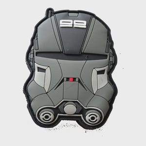 Pode incluir: Um patch cinza e preto em forma de capacete com o número "99" no topo. O patch tem um detalhe quadrado vermelho e duas características circulares na parte inferior. O patch é provavelmente um item colecionável ou decorativo.