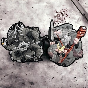 Könnte beinhalten: Zwei gestickte Patches, einer zeigt ein graues und schwarzes Monster mit scharfen Krallen und Zähnen, der andere einen grauen und weißen Zauberer, der einen roten Stab und ein gelbes Objekt hält.