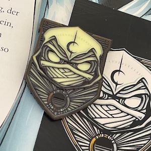 Könnte beinhalten: Schildförmiger Emaille-Pin mit stilisiertem Charakterdesign, cremefarbenem Gesicht und einem Halbmondsymbol. Der Pin hat einen dunkelbraunen Rand und ein Sonnenstrahl-Design am unteren Rand. Der Pin liegt auf einer schwarzen Oberfläche.