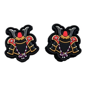 Op de afbeelding: Twee zwarte schoenbedels met een gedetailleerd ontwerp van een samoeraihelm. De helmen hebben rode, gouden en zilveren accenten, met een zwarte basis en een paarse en witte kralenketting.