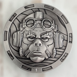 May include: A round, silver-colored metal medallion featuring a stylized face with goggles and ear coverings. The face has a stern expression. The number "1" is at the bottom. The medallion has an outer decorative border.