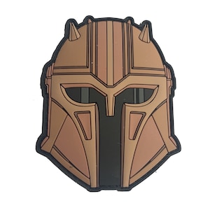 May include: A brown and black patch featuring a stylized helmet design. The patch has a detailed, geometric pattern with a black border. The helmet has two small horns on top and a visor.