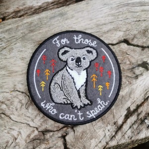 以下が含まれることがあります： 中央に座っているコアラが描かれた灰色の刺繍パッチ。コアラの周りはオレンジと黄色の小さな木々で囲まれています。「For those who can't speak.」という文字がコアラの周りに書かれています。