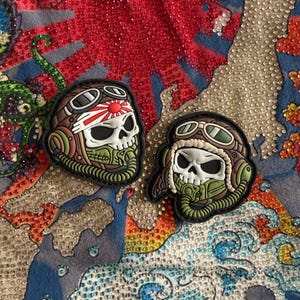 May include: Two skull-shaped patches. One patch features a skull wearing a brown aviator helmet with goggles and a rising sun emblem. The other patch has a similar design, with a brown aviator helmet and goggles. Both have green and black accents.