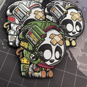 Peut inclure: Écussons brodés avec un motif de panda dans un style futuriste et militaire. Les écussons comportent le texte "7SKETCHES UNIVERSE-WIDE" et "出陸". Le panda est représenté dans différentes tenues, comme une combinaison spatiale et un équipement militaire.