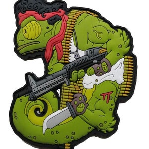 Puede incluir: Un camaleón verde de dibujos animados que lleva un pañuelo rojo y sostiene una ametralladora y un cuchillo. El camaleón lleva un tanque blanco y tiene un cinturón de balas alrededor del cuerpo.