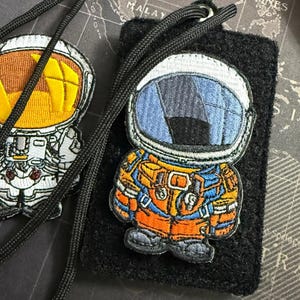 Op de afbeelding: Geborduurde astronautenpatch in oranje, blauw en wit, met een zwart koord. De patch zit op een zwarte vilt achtergrond. De helm van de astronaut heeft een blauw vizier. Het pak van de astronaut is oranje en blauw.