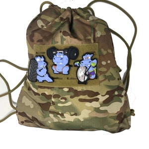 Puede incluir: Mochila con cordón de camuflaje con tres parches de hipopótamos de dibujos animados. Los parches representan hipopótamos levantando pesas, sosteniendo un neumático y usando una máquina de pesas. La bolsa tiene un cierre de cordón y cordones verde oliva.