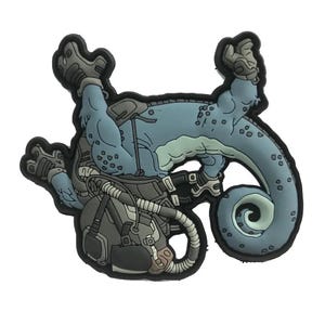 May include: A blue and gray cartoon-style patch featuring a reptilian creature in a spacesuit. The creature has a long tail and is wearing a helmet and backpack. The patch has a black border and is likely made of rubber or a similar material.
