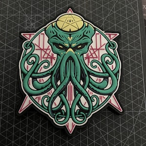 Op de afbeelding: Een geborduurde patch met een gestileerde octopus in groen en geel, met een derde oog op zijn voorhoofd. De octopus wordt omringd door een witte cirkel met een rood en wit geometrisch patroon.