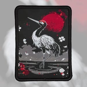 May include: A rectangular embroidered patch featuring a white crane standing in water. The design includes a large red sun in the background, scattered white cherry blossom petals, and a dark black border surrounding the entire rectangular shape.
