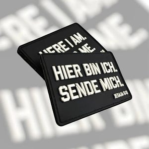 May include: Two black rectangular patches with white raised lettering. One patch reads "HERE I AM. SEND ME." The other patch reads "HIER BIN ICH. SENDE MICH." with "JESAJA 6:8" below. The patches are slightly overlapping.