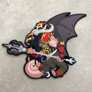 以下が含まれることがあります： 黒いひげを生やし、三叉の槍を持った角のある悪魔が描かれたカラフルなパッチ。悪魔はコウモリの羽、赤と金のアクセント、丸まった尾を持っています。パッチはニュートラルグレーの表面にあります。