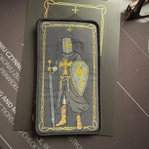 Peut inclure: Patch brodé représentant un chevalier en armure tenant une épée et un bouclier. Le chevalier porte un casque avec une croix. Le patch est noir avec des accents dorés.