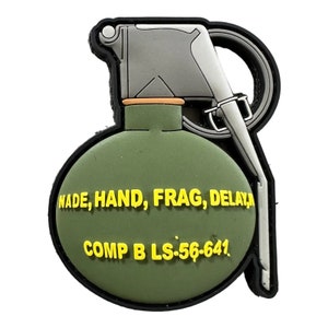 Puede incluir: Un parche verde y gris con forma de granada con texto amarillo que dice "NADE, HAND, FRAG, DELAY" y "COMP B LS-56-641." El parche tiene un borde negro y un asa plateada.