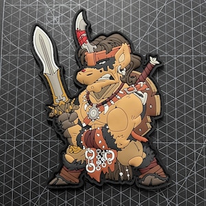 Könnte beinhalten: Ein Cartoon-Patch mit einem Krieger mit brauner Haut, braunen Haaren und einem entschlossenen Gesichtsausdruck. Der Krieger hält ein großes Schwert und ein kleineres Messer und trägt Rüstung und Accessoires. Der Patch hat einen schwarzen Rand.