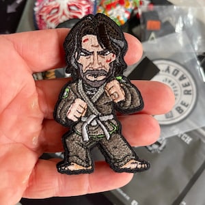 Könnte beinhalten: Gestickter Aufnäher mit einer Comicfigur in einem schwarzen Gi mit einem weißen Gürtel, grünen Akzenten und einem schwarzen Bart. Die Figur hat einen ernsten Gesichtsausdruck und befindet sich in Kampfstellung.