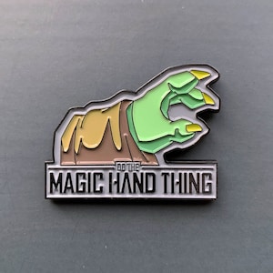 Könnte beinhalten: Emaille-Anstecknadel mit einer grünen Hand mit gelben Fingernägeln, die aus einem braunen und beigen Ärmel kommt. Die Nadel hat einen schwarzen Rand und den Text "DO THE MAGIC HAND THING" in Weiß.
