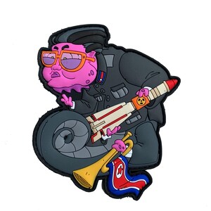Può includere: Una toppa a fumetti raffigurante un personaggio dalla pelle rosa in uniforme militare grigio scuro e occhiali arancioni. Il personaggio tiene in mano un razzo con un simbolo nucleare e una tromba d'oro con una bandiera nordcoreana.