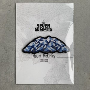 May include: A patch featuring a mountain range design in shades of blue and white, with the text "Mount McKinley" and "136/300" below. The patch is set against a white background with a topographic map design, and the text "THE SEVEN SUMMITS" is at the top.