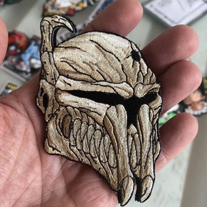 May include: A beige and black embroidered patch of a creature with a skull-like face and sharp teeth. The creature has a large, pointed head and a pair of horns.