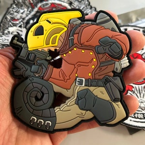 Puede incluir: Un parche colorido que presenta a un soldado de dibujos animados con un casco amarillo y una chaqueta marrón, sosteniendo un arma. El parche tiene un borde negro y está sobre un fondo borroso de otros parches.