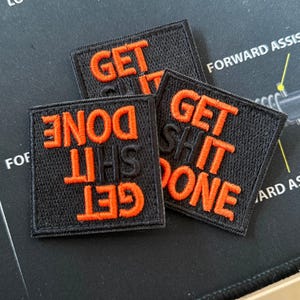 Puede incluir: Tres parches de tela negra con texto bordado en naranja. Los parches dicen "Get Shit Done".