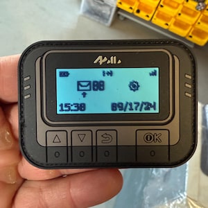 May include: A black and grey device with a blue screen displaying the time 15:38, the date 89/17/24, and icons for email, a clock, and a cog. The device has four buttons labelled "▲", "▼", "↺", and "OK".