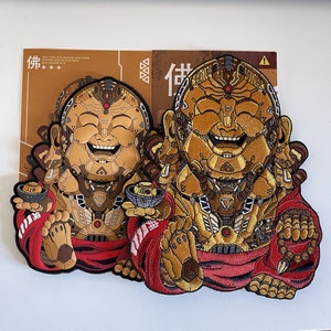 May include: Two embroidered patches featuring a smiling Buddha with a robotic design. The patches are primarily gold and brown, with red accents on the robes and a small bowl. The background card has text in English and Asian characters.