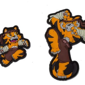 Puede incluir: Dos parches bordados para planchar que representan a un tigre de dibujos animados en una pose de lucha. El parche más grande muestra al tigre con un vendaje en el brazo y una expresión decidida. El parche más pequeño muestra al tigre en una pose más juguetona con la lengua fuera.