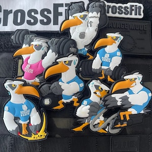 May include: A set of eight colourful, cartoon seagull patches with the text "Battle of the Beach" on each. The seagulls are depicted lifting weights, with some wearing blue, pink and white shirts. The patches are made of a rubber-like material and have a textured surface.