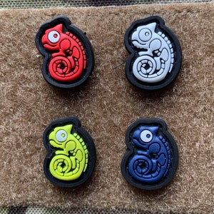 May include: Four colorful chameleon-shaped patches. The patches are red, white, yellow-green, and blue, each with a black border. The chameleons have detailed designs and are set against a tan background.