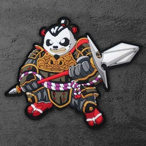 Op de afbeelding: Een kleurrijke patch met een panda-krijger in gedetailleerde bepantsering, met een speer. Het ontwerp bevat rood, goud, zwart en wit, met een paars en wit touw. De panda heeft een serieuze uitdrukking.