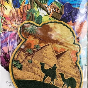 Op de afbeelding: Een geborduurde patch in geel, groen en bruin die een woestijnscène voorstelt met twee kamelen die voor de piramides lopen. De patch heeft een gele rand en is omgeven door een doorzichtige plastic zak.