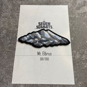 May include: Embroidered patch featuring a mountain range design with the text "The Seven Summits" and "Mt. Elbrus". The patch is black, gray, and blue, with a black border. The card reads "001/300".