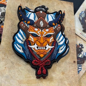 May include: A blue, white and orange samurai mask with a red rope tied around the neck. The mask has a fierce expression with sharp teeth and horns. The mask is made of a textured material and has a 3D effect.