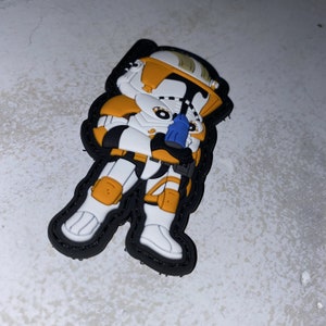 Op de afbeelding: Een witte en oranje cartoon kloon soldaat met een blauwe blastergeweer. De soldaat draagt een helm en heeft een zwarte omtrek.