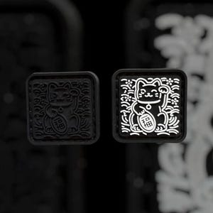 May include: Two square patches featuring a raised relief design of a beckoning cat holding a coin with a character. One patch is solid black, while the other has a black base with white outlines highlighting the cat, floral patterns, and coin.