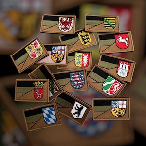 Op de afbeelding: Een verzameling geborduurde patches met verschillende heraldische wapens en emblemen. Elke patch heeft een rechthoekige vorm met een bruine rand, een zwarte en olijfgroene streep en een kleurrijk wapenontwerp. De patches zijn waarschijnlijk voor kleding of accessoires.