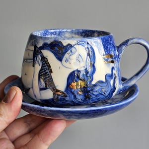 May include: A blue and white ceramic teacup and saucer set. The cup features a hand-painted design of a figure with long hair and a fish, with gold accents. The saucer is a matching blue. The cup has a rounded shape with a handle.