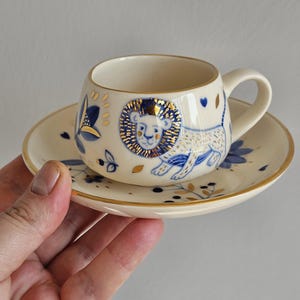 Könnte beinhalten: Cremefarbenes Keramik-Tee-Set. Die Tasse hat ein blaues und goldenes Löwen-Design mit blauen Blumen- und Goldakzenten. Die Untertasse hat passende Blumen- und Golddetails. Das Set wird in einer Hand gehalten.