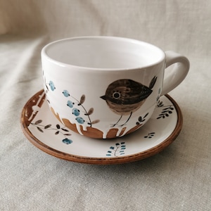Könnte beinhalten: Eine weiße Teetasse und Untertasse aus Keramik mit einem Design aus braunem Vogel und blauen Beeren. Die Tasse hat einen braunen Tropfeffekt am Rand.