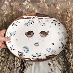 May include: White ceramic oval serving platter with a brown rim. The platter is decorated with hand-painted blue and brown floral designs, two birds, and a nest with eggs.