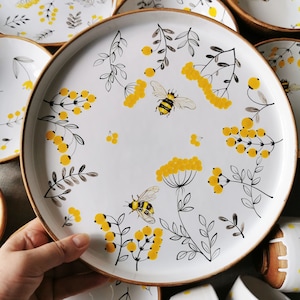Könnte beinhalten: Weiße Keramikplatte mit einem schwarzen und gelben Blumendesign mit Bienen und Beeren. Die Platte hat einen braunen Rand.
