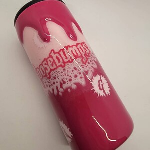 May include: A pink and white tumbler with a black lid. The tumbler has a white design with the words "Goosebumps" and a "G" with a white splatter effect.